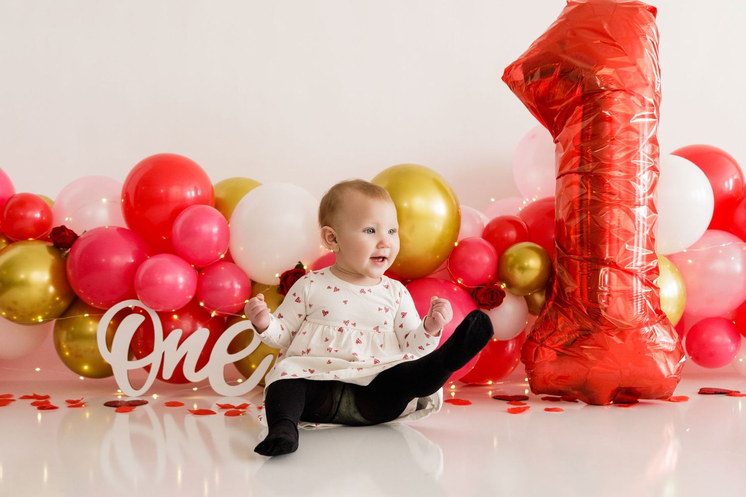A baby in black pants crawls across a pink and red balloon-decorated backdrop during a first birthday photoshoot.
