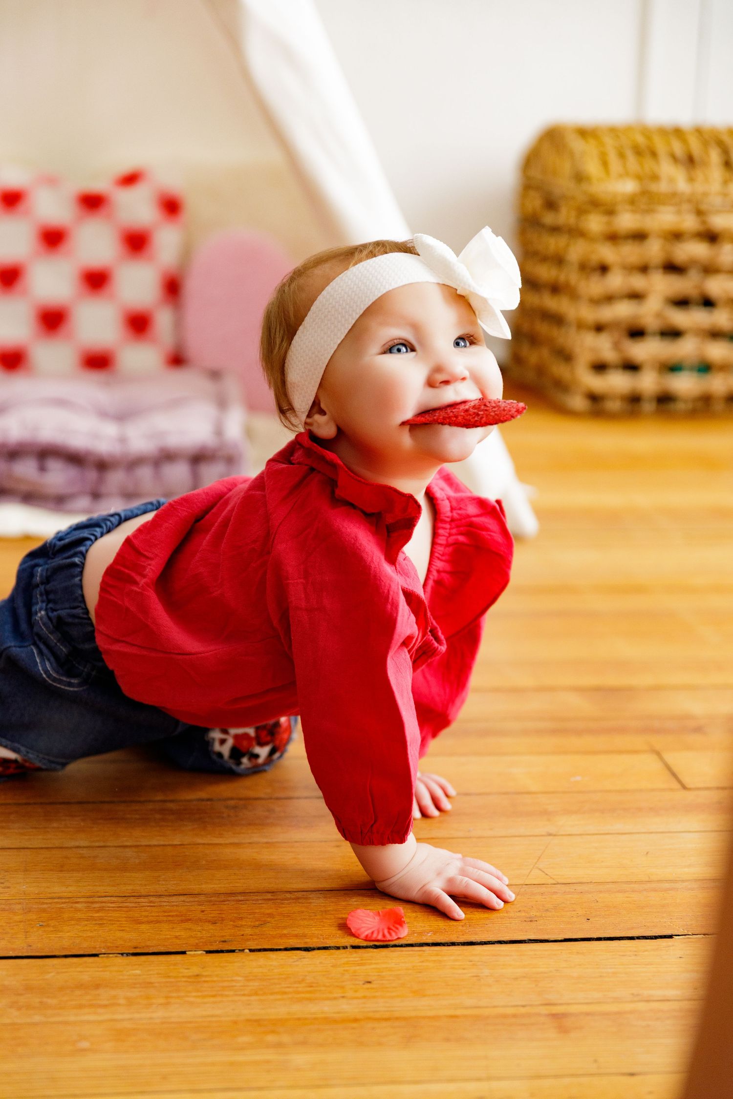 Baby in red sweater and white headband crawling on hardwood floor with playful expression.