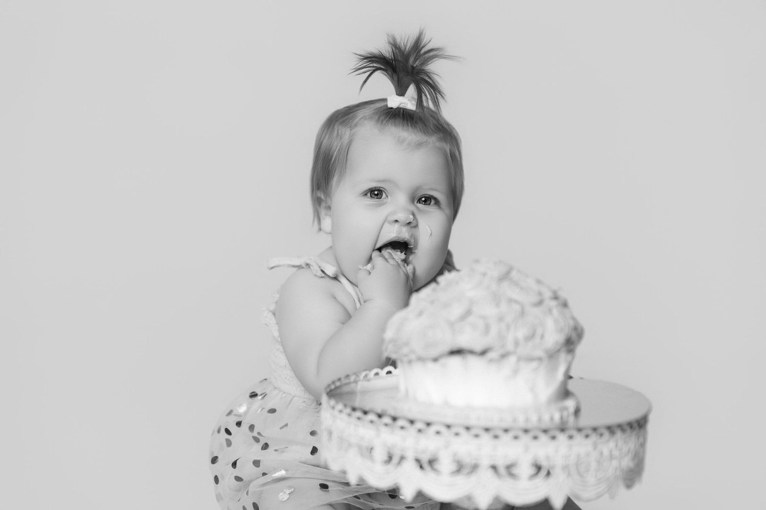 Baby reaching for pink frosted cake on ornate white pedestal stand.