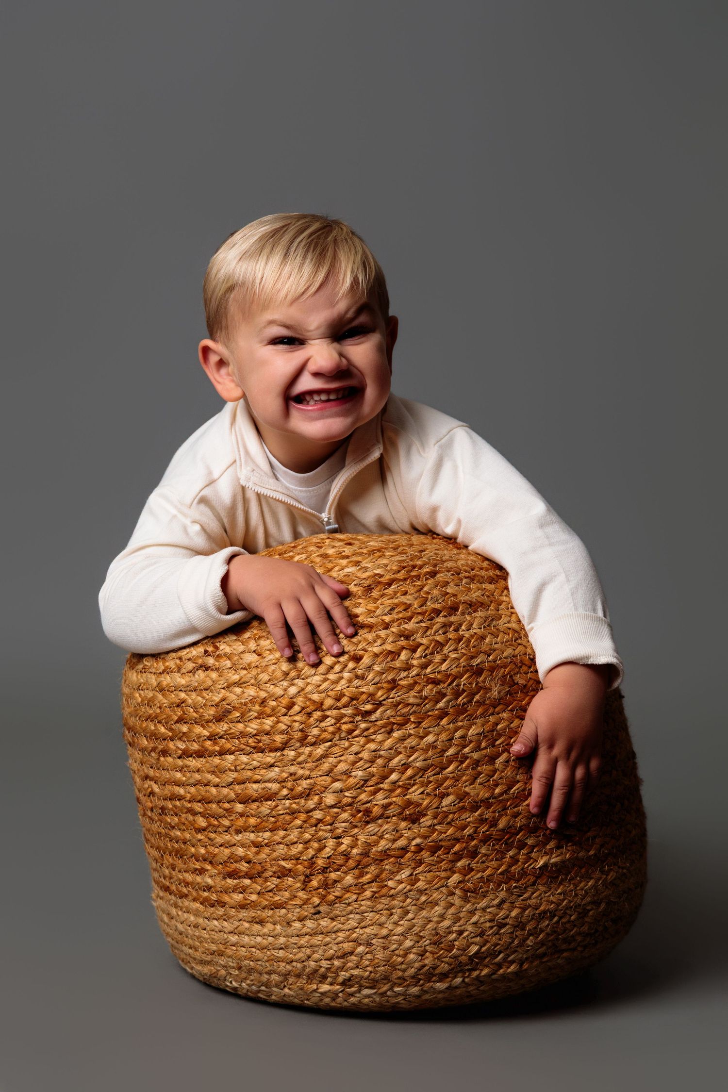 A child plays with a large woven straw ottoman cushion against a gray studio backdrop.