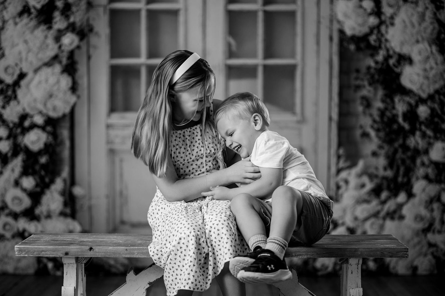 Black and white photo of a girl and young boy sitting on a bench, sharing a tender sibling moment.