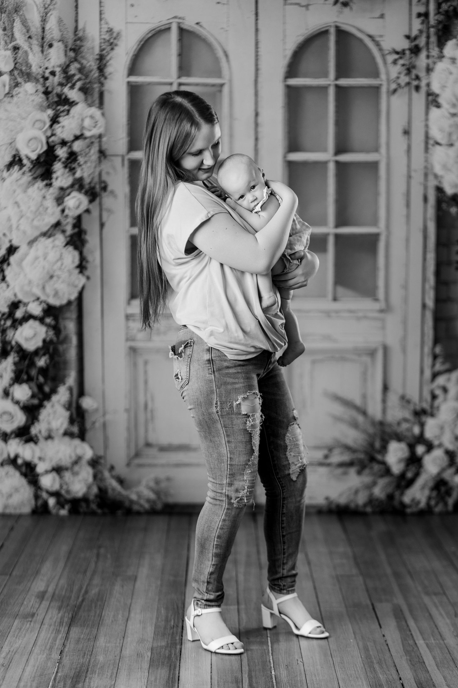 Black and white photo of a young mother tenderly holding her newborn baby while standing in front of a floral backdrop.