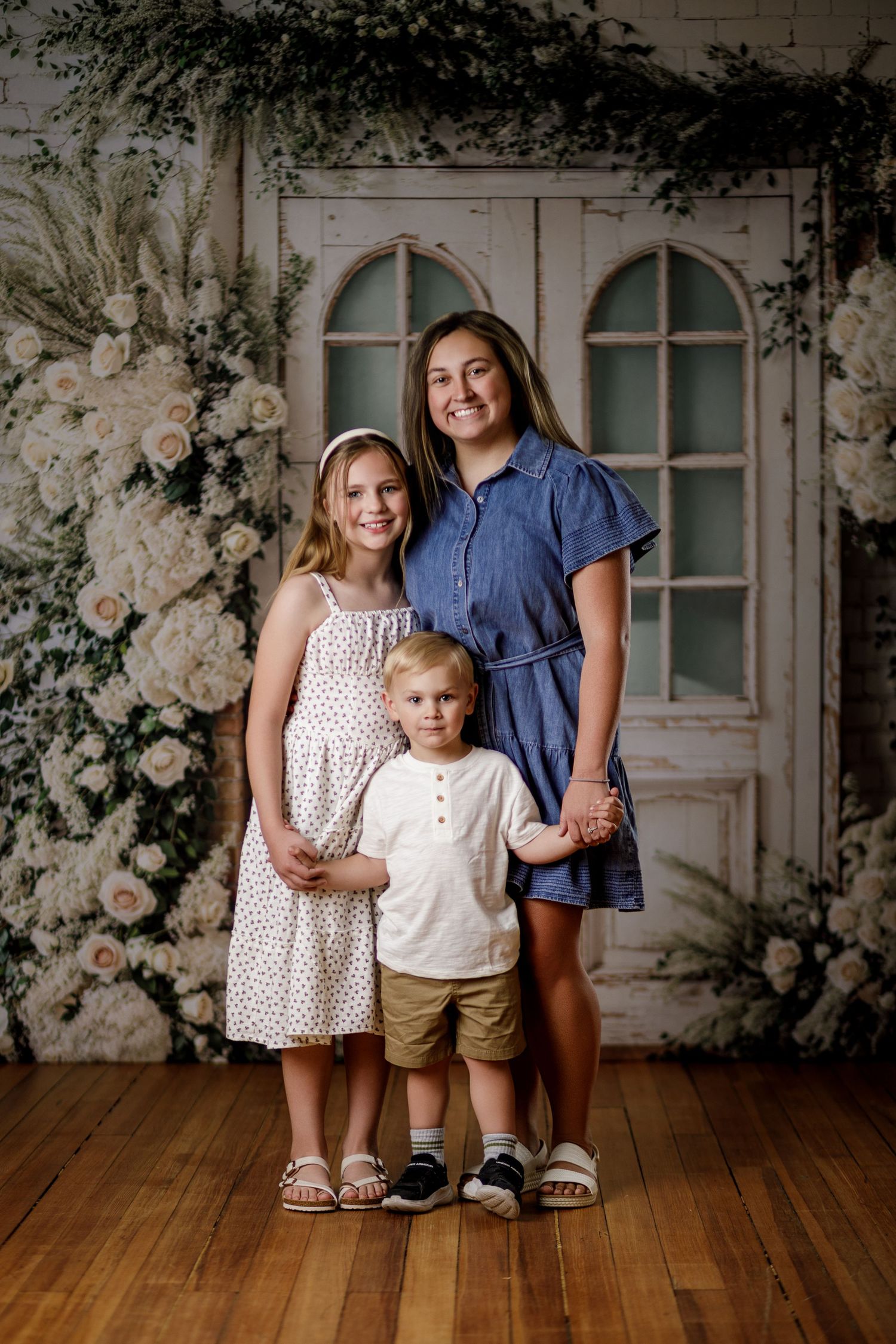 Three children pose together in front of a floral backdrop with a vintage white door for a professional portrait.