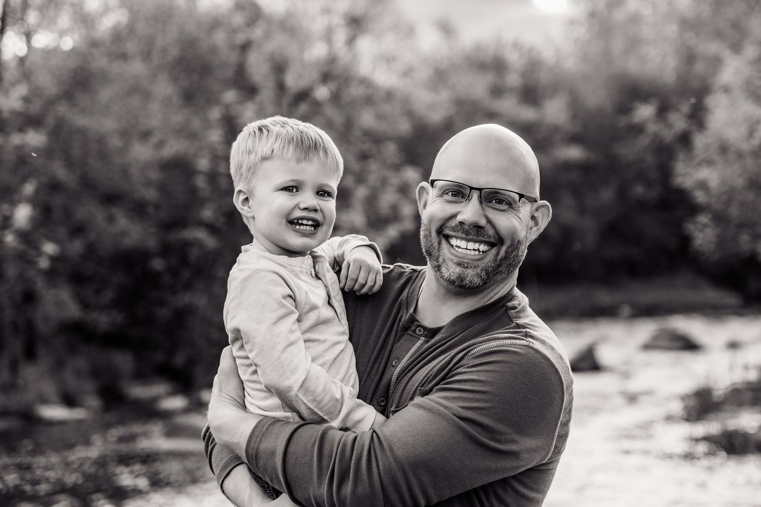 Bald man with glasses smiling and holding a laughing toddler boy outdoors in a black and white photo.