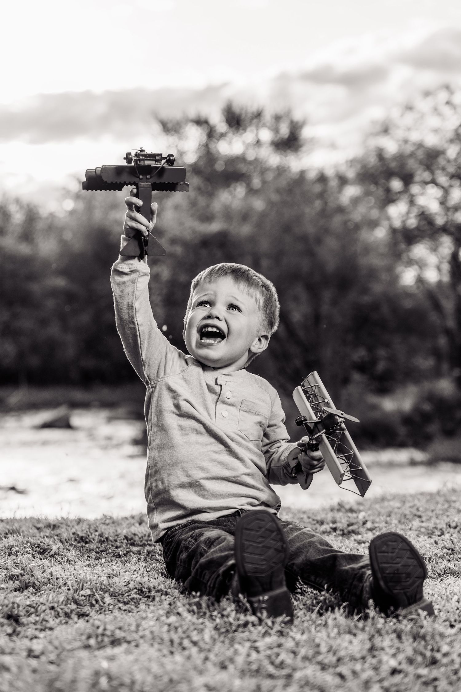 Joyful toddler boy sitting on grass playing with wooden toy airplanes outdoors in black and white photo.