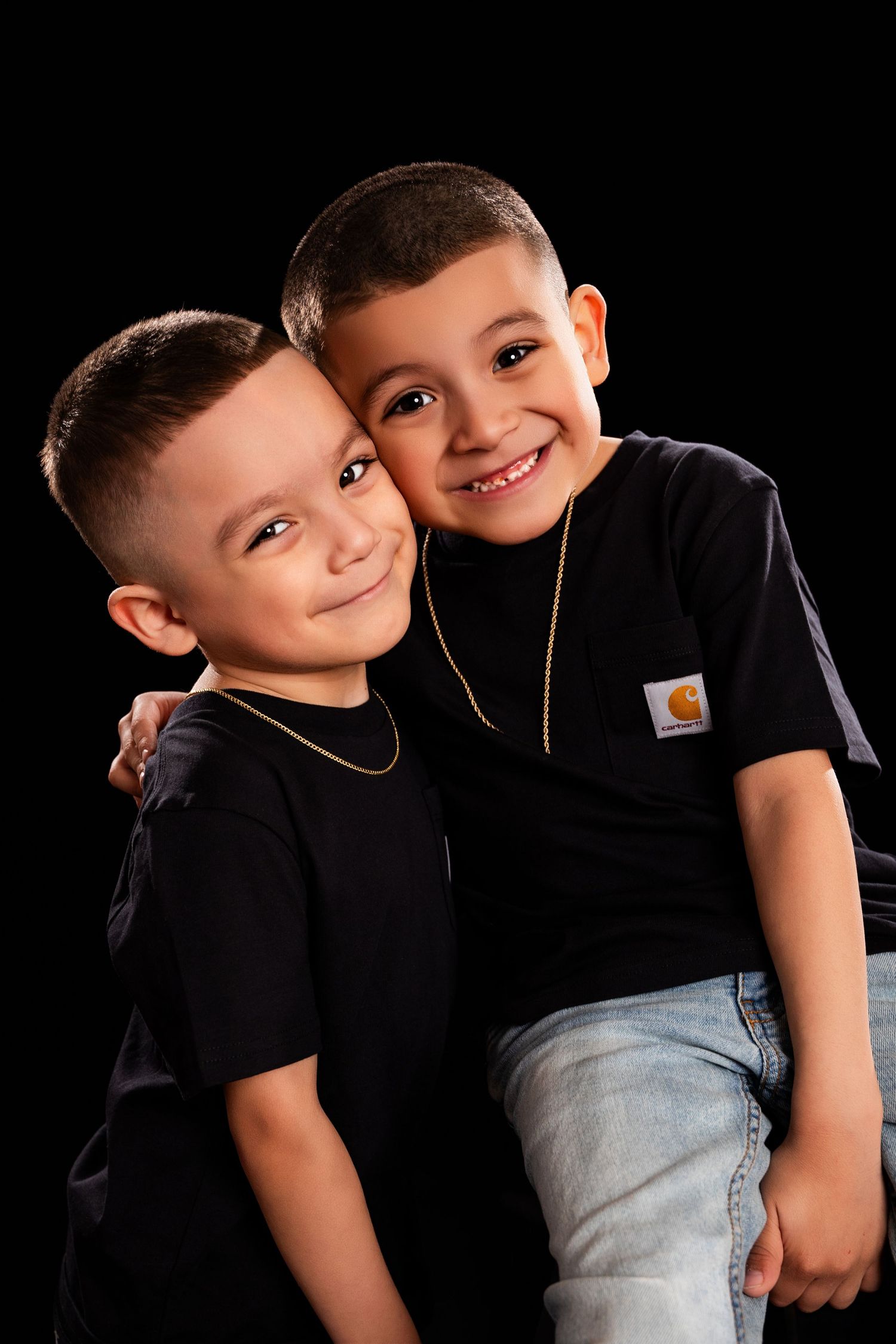 Two children wearing matching black t-shirts and gold necklaces pose together against a dark background for a portrait.
