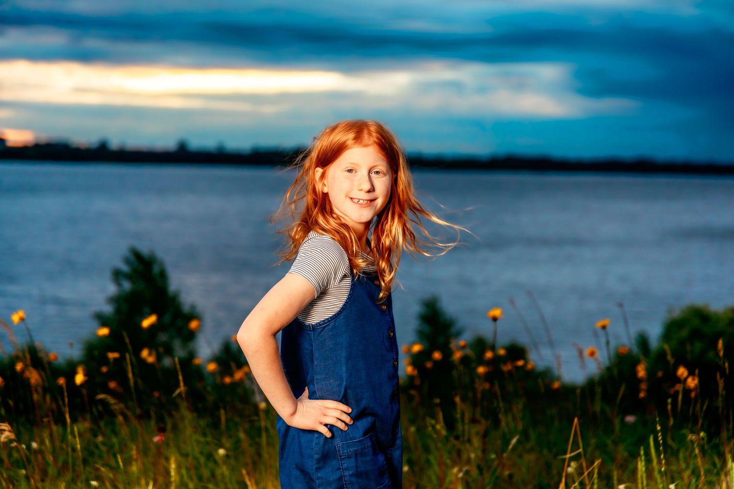 Young people posing in a field of wildflowers by a lake with dramatic blue sky overhead.