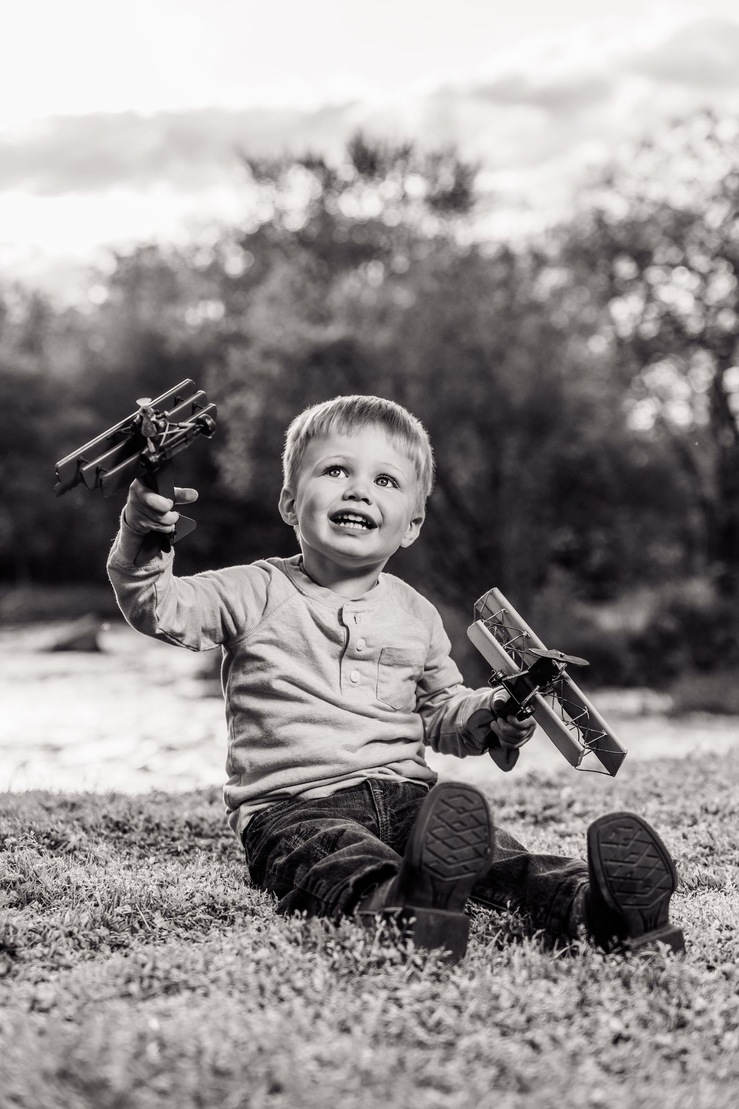Black and white photo of a joyful toddler boy sitting on grass, holding two toy wooden airplanes outdoors.