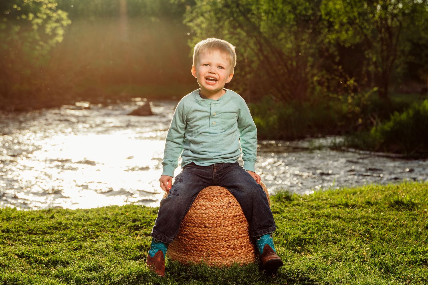 A laughing toddler boy sits on a wicker basket outdoors by a sunlit river surrounded by lush green grass.