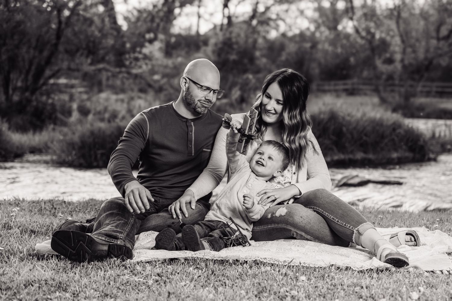 Black and white family photo of parents and baby sitting on blanket outdoors near trees.