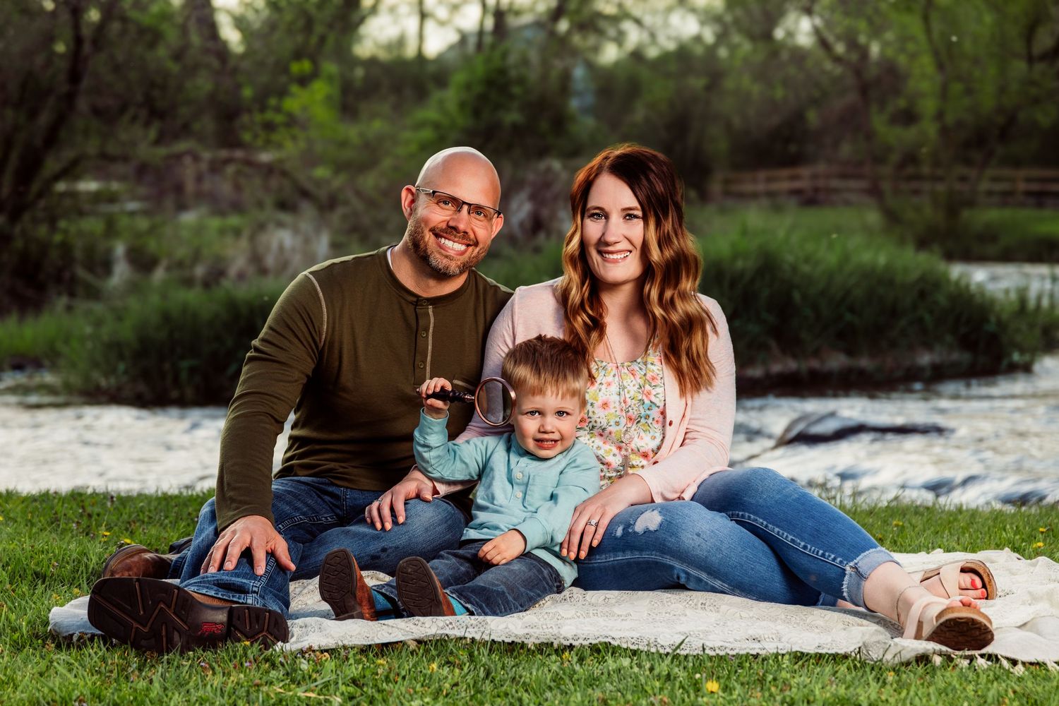 Happy family of three smiling together on a blanket outdoors near a river on a sunny day.