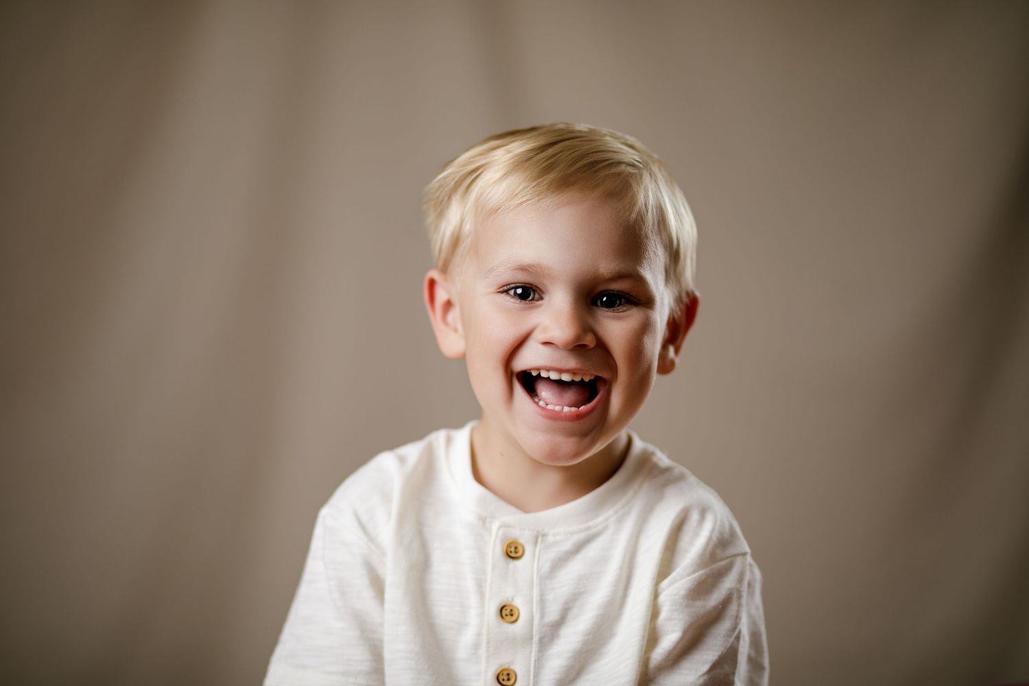 Laughing blonde toddler boy in white henley shirt poses for portrait against neutral beige background.