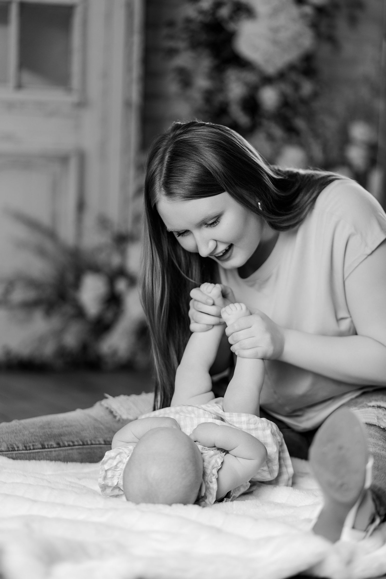 Black and white photo of a laughing mother playing with her baby's feet on a soft blanket.