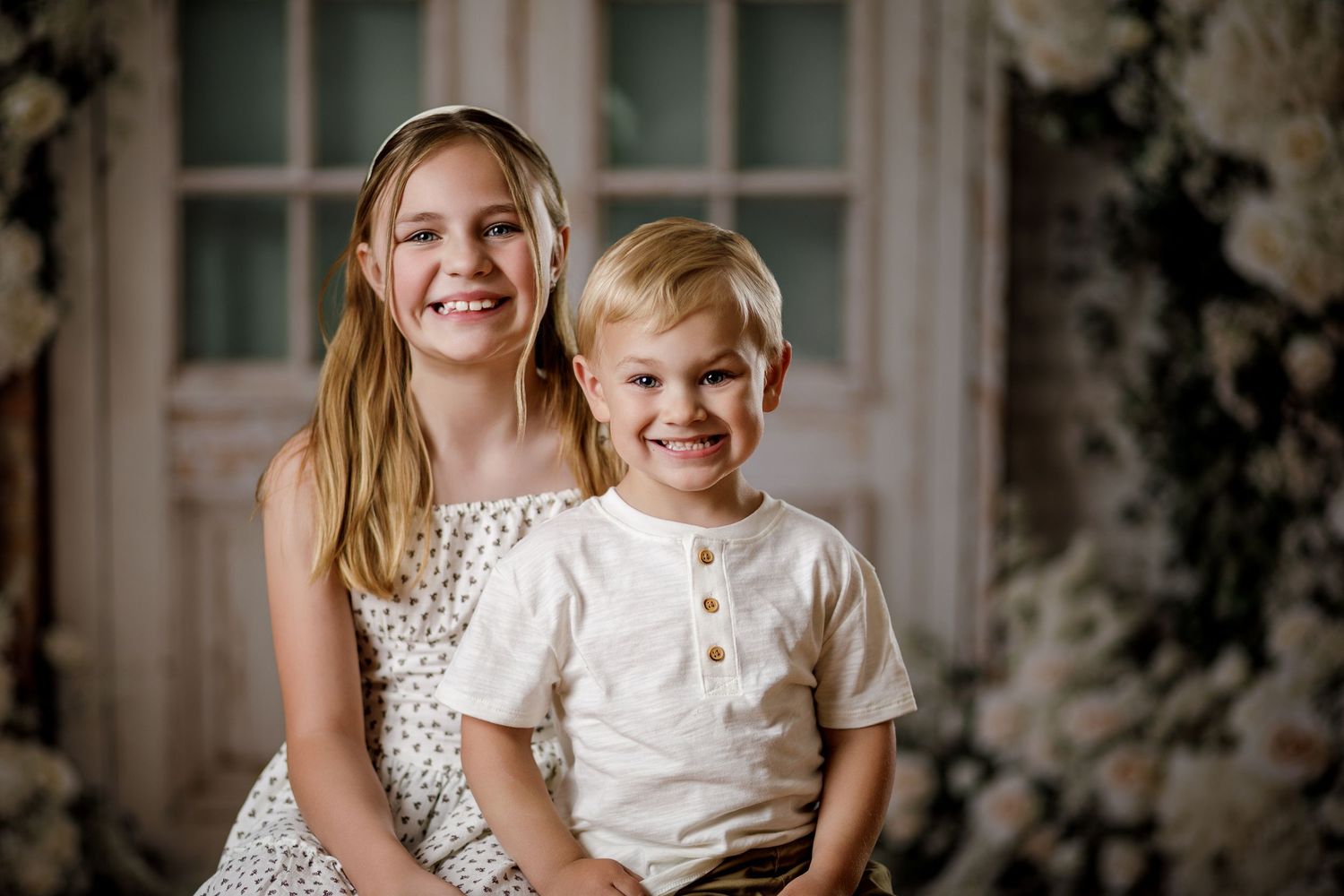 Smiling girl and boy siblings pose together in front of a floral backdrop in a cheerful portrait.