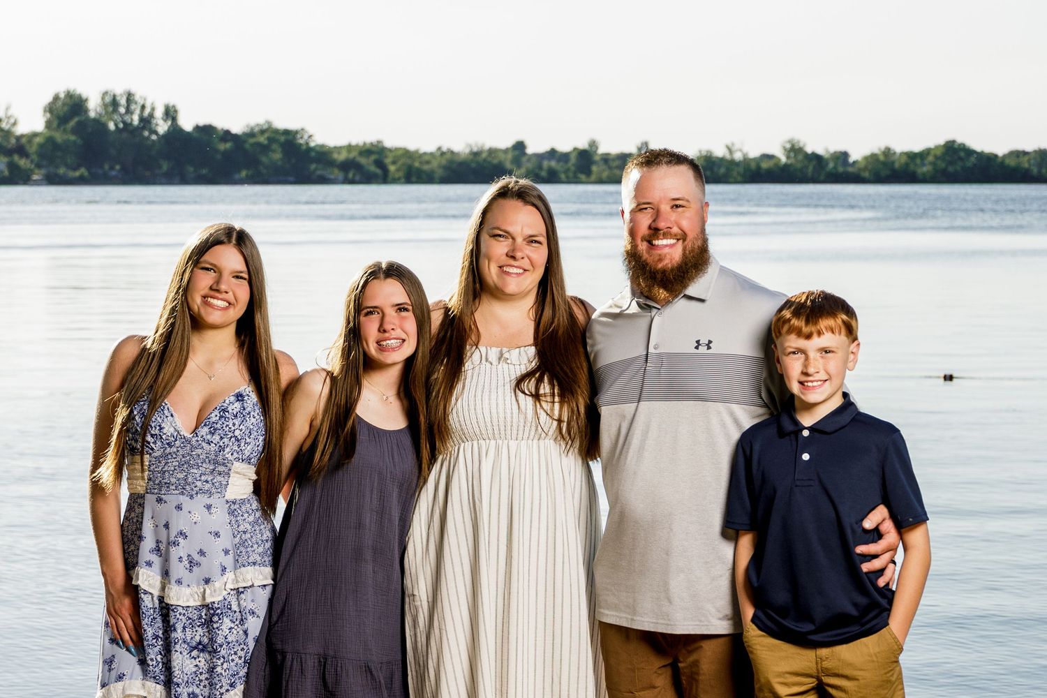 Family of five smiling together by a lake at sunset, with parents and three children posing on a waterfront.