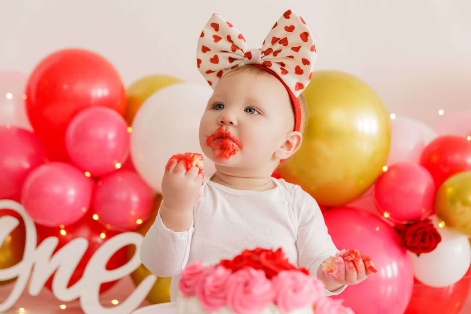 Joyful baby in red and white bow surrounded by pink and gold balloons at cake smash photography session.