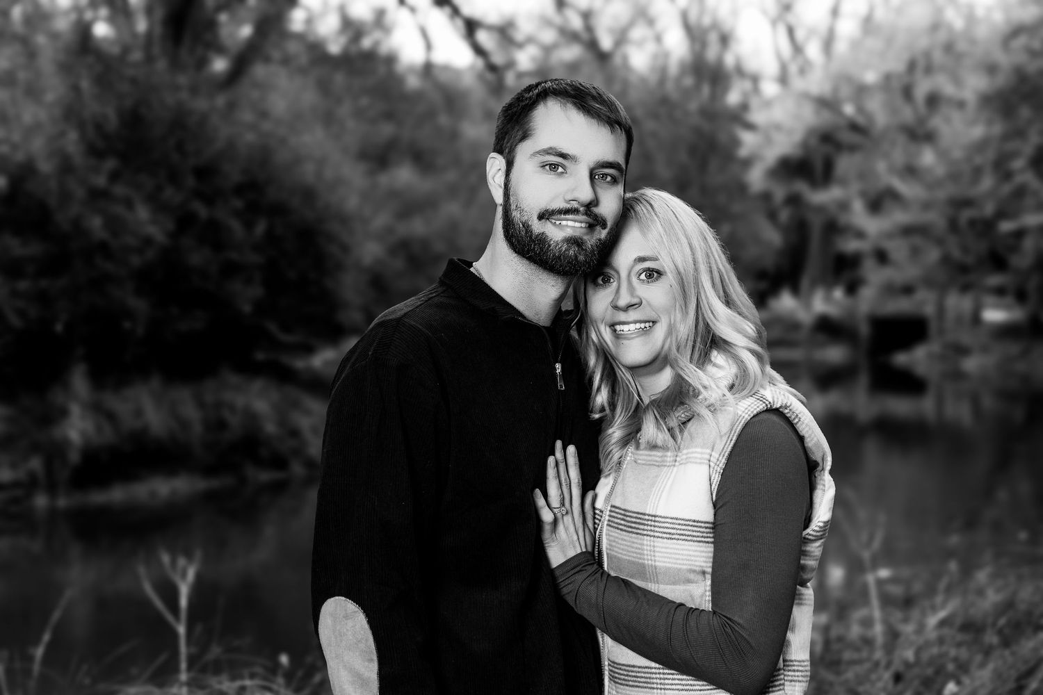 Romantic engagement photo of couple laughing together by lake in fall setting.