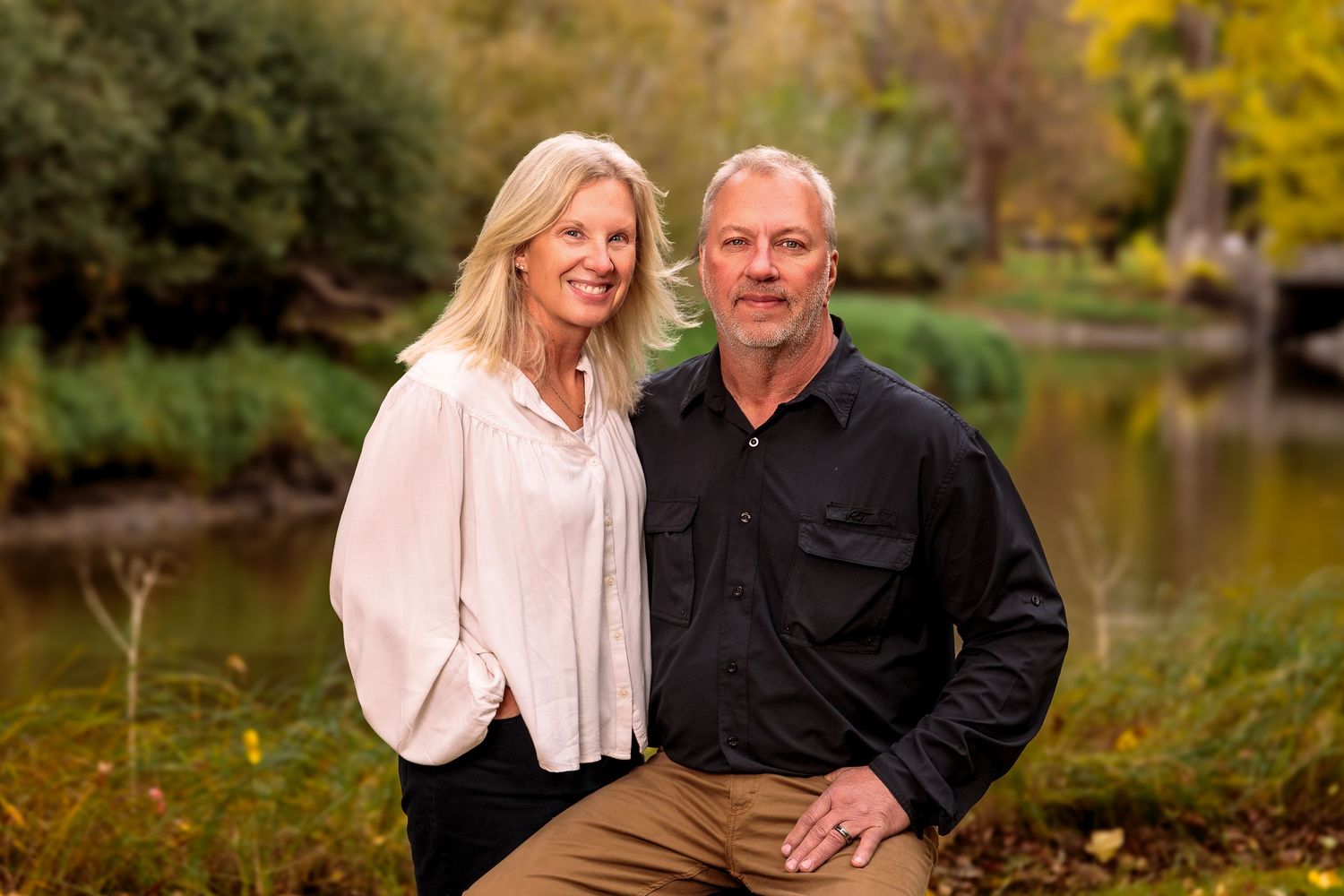 Couple sitting together outdoors by a pond surrounded by fall foliage.