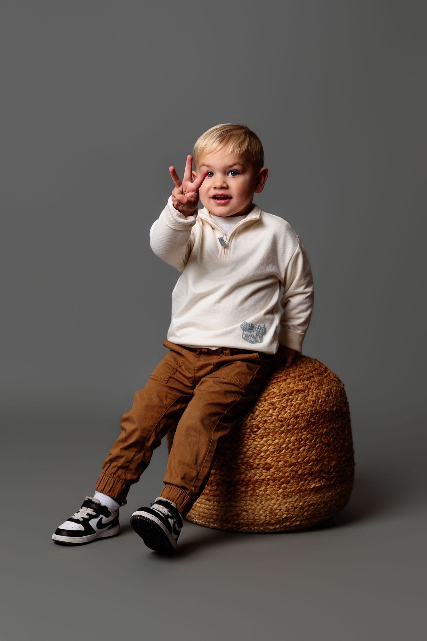 Young child sitting on brown textured ottoman wearing white sweater and brown pants against grey backdrop.