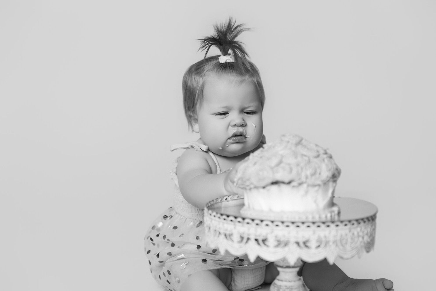 Baby reaching for white cake on decorative stand during first birthday photoshoot.