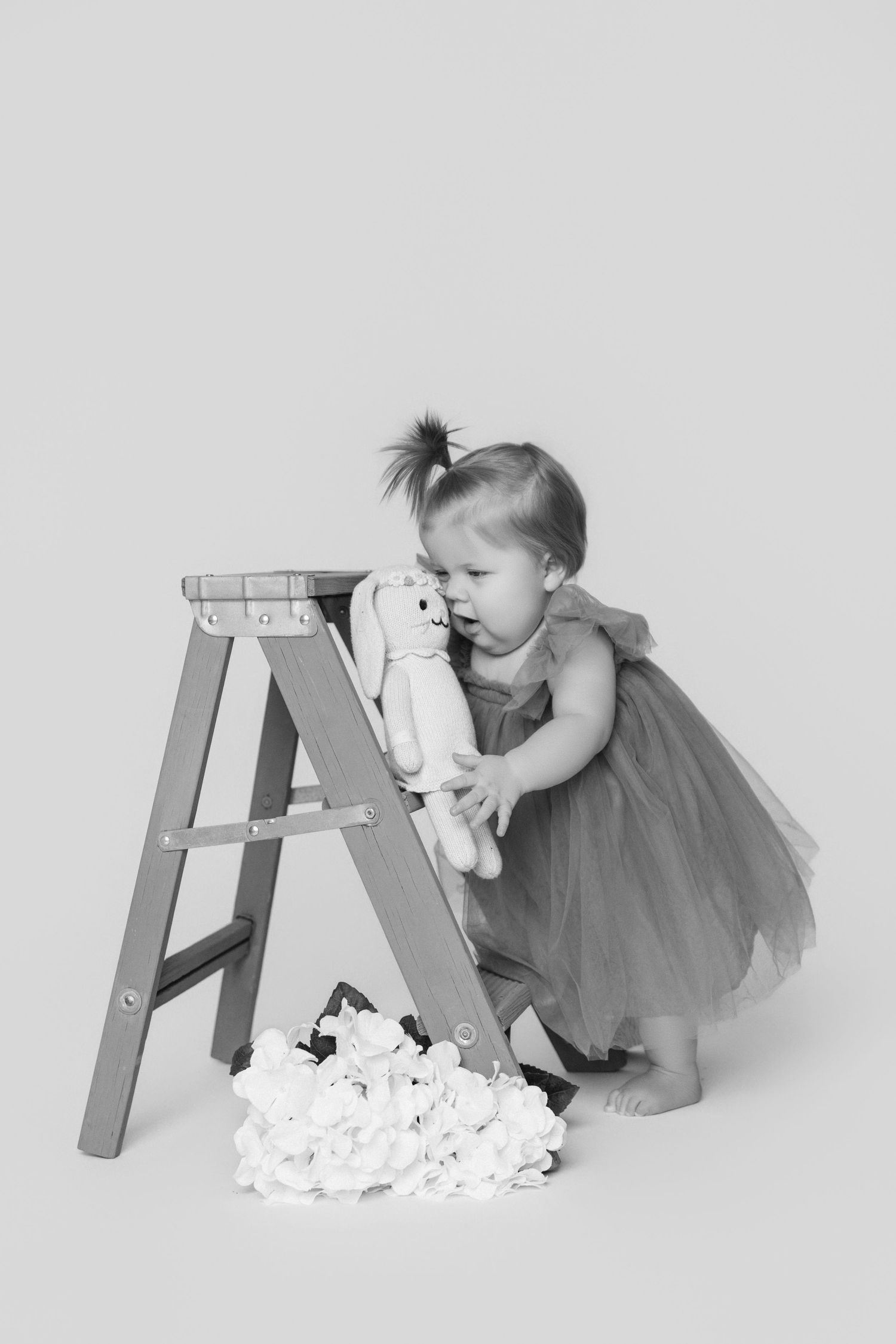 Baby in gray tulle dress explores wooden ladder prop with white fabric.
