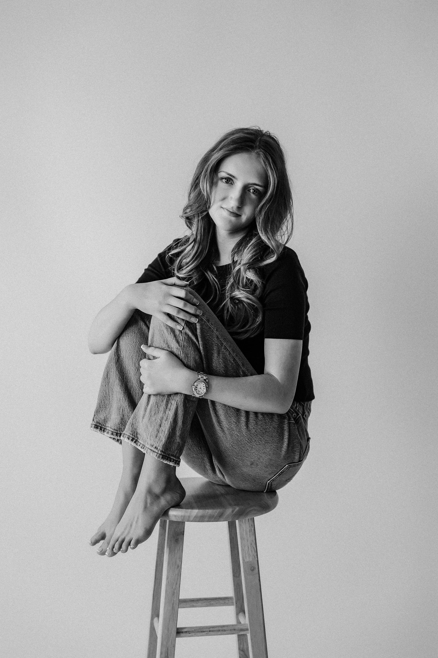 Black and white portrait of a young woman sitting on a wooden stool, hugging her knees, wearing jeans and a black top.