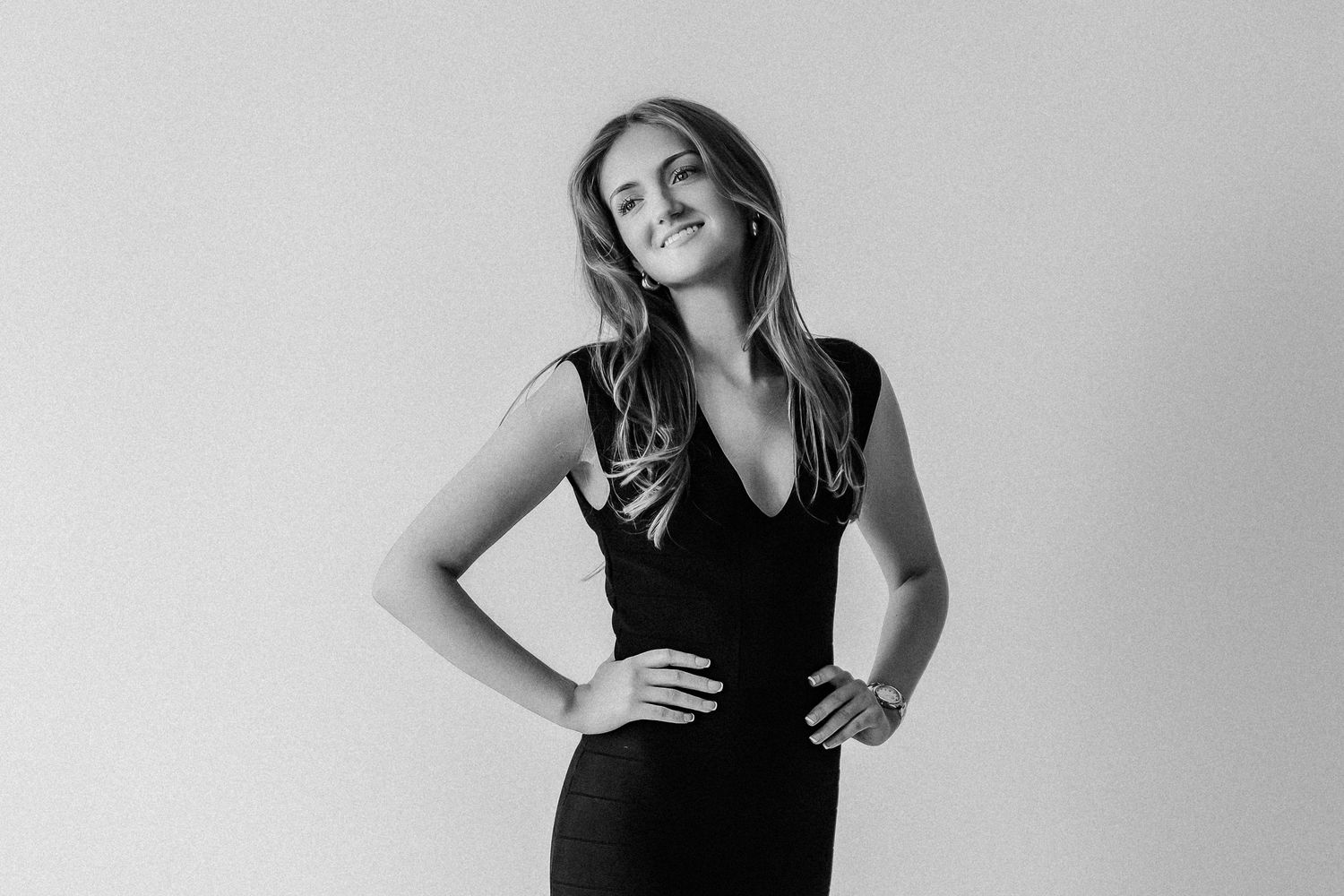 Black and white studio portrait of confident young woman in sleeveless black dress with hands on hips.