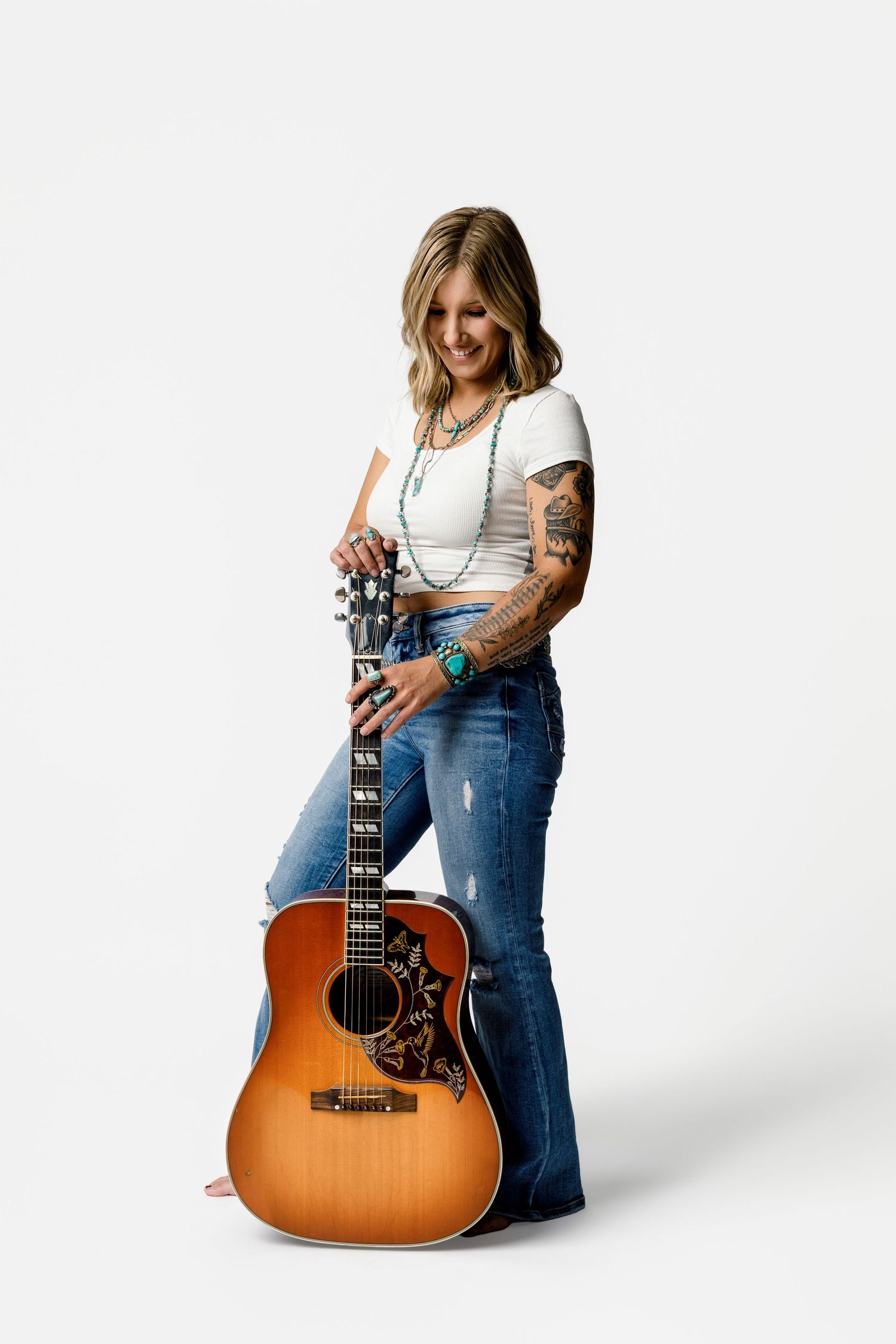 Tattooed female musician posing with acoustic guitar in white top and jeans against white background.