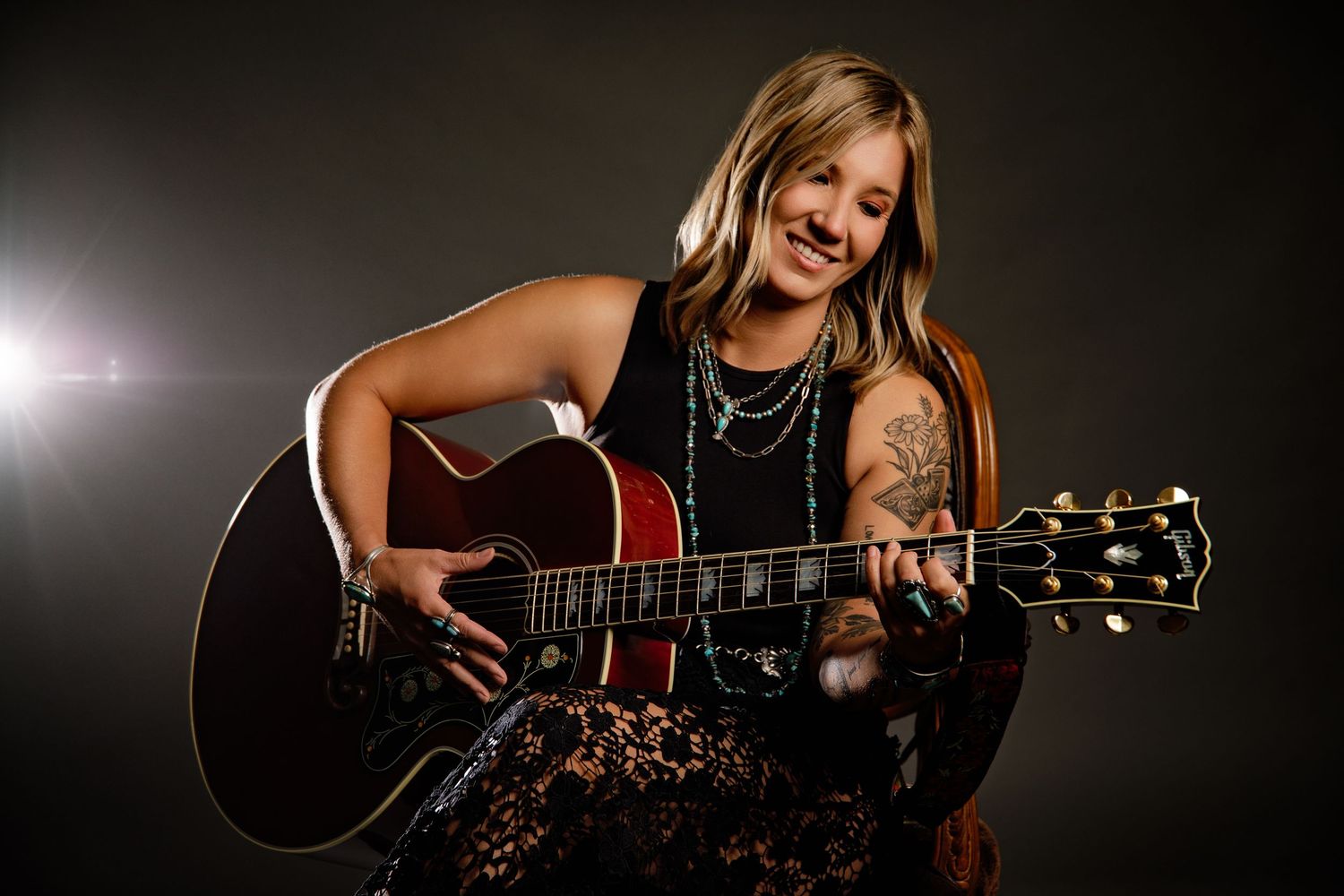 Smiling woman with tattoos playing acoustic guitar on stage with turquoise necklace.