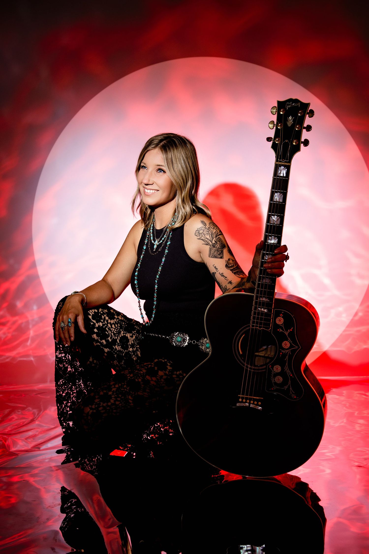 Tattooed woman sitting with black acoustic guitar against vibrant red atmospheric background.