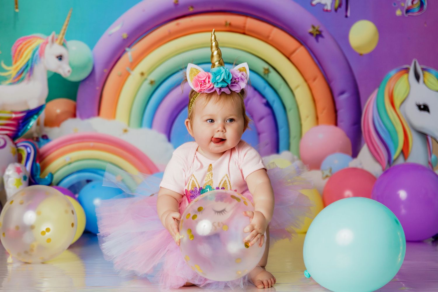 Baby girl in unicorn costume with horn headband plays with balloons among colorful rainbow and unicorn party decorations.