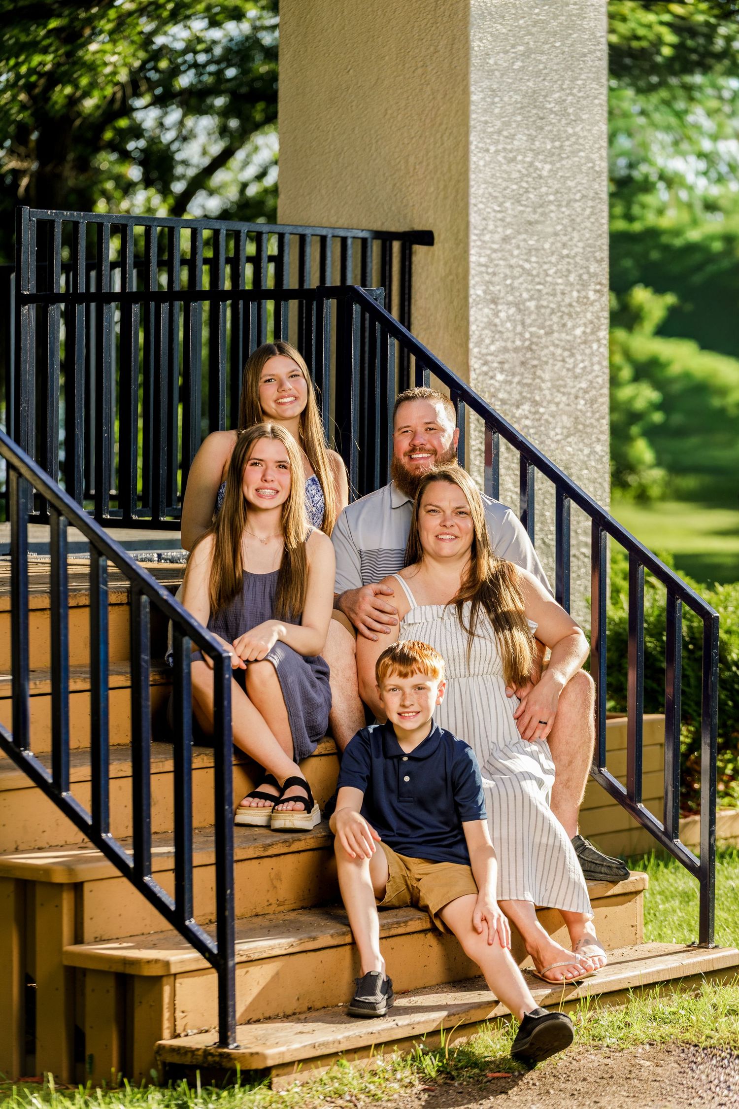 Family of five sitting on outdoor steps by black iron railings, smiling together in warm golden light.
