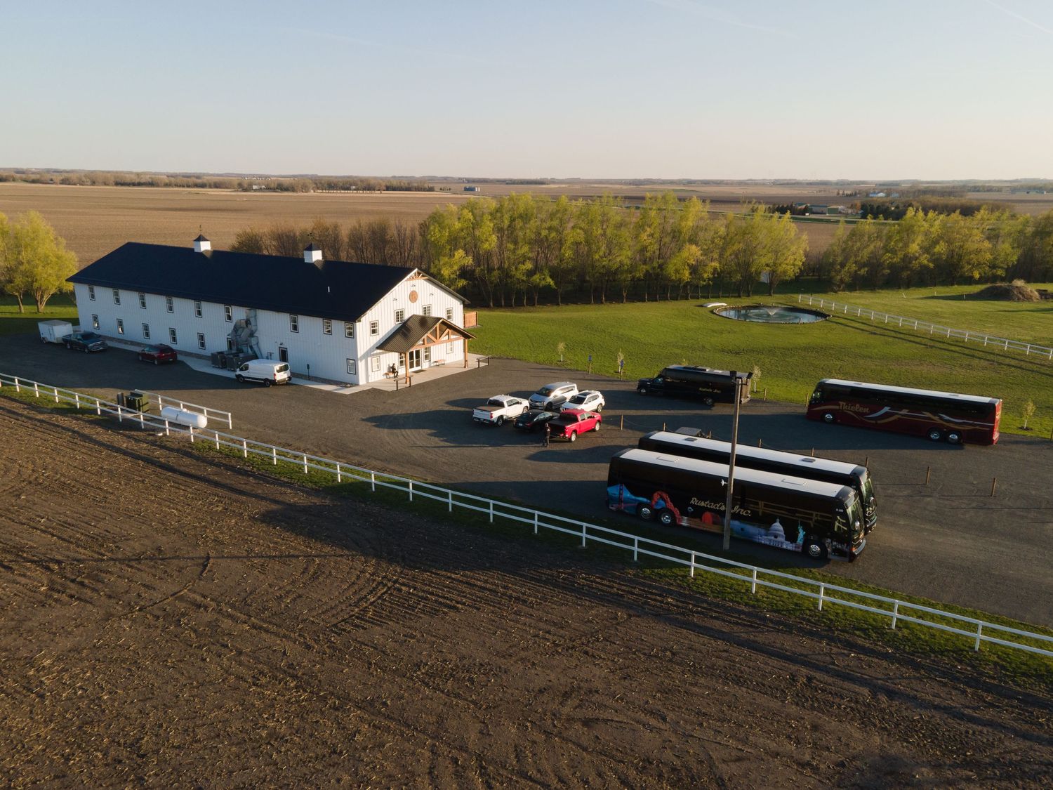 Aerial view of a white farmhouse with white fencing and dirt field surrounded by parked vehicles and trailers.