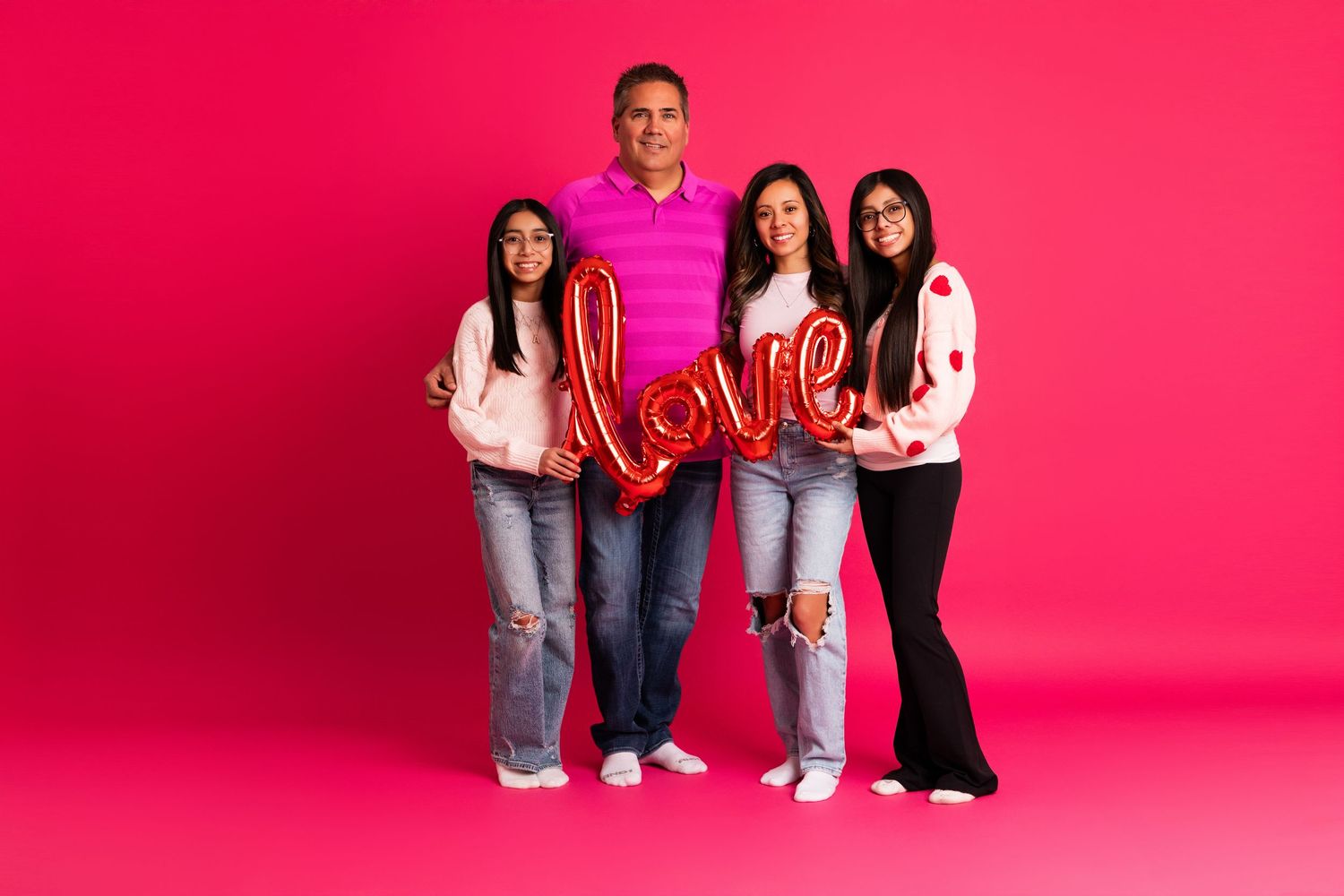 Family of four posing with a red love balloon script against a vibrant pink background for Valentines Day.