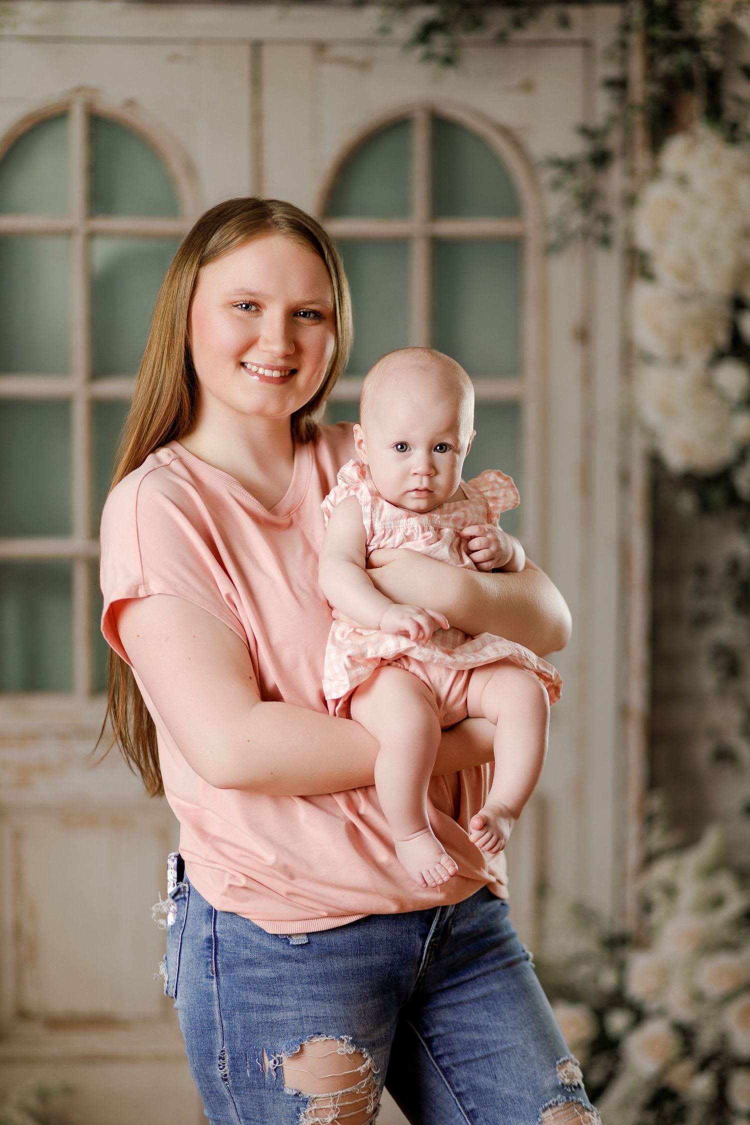 Smiling young mother in pink t-shirt and jeans holds her baby girl in a floral studio setting.