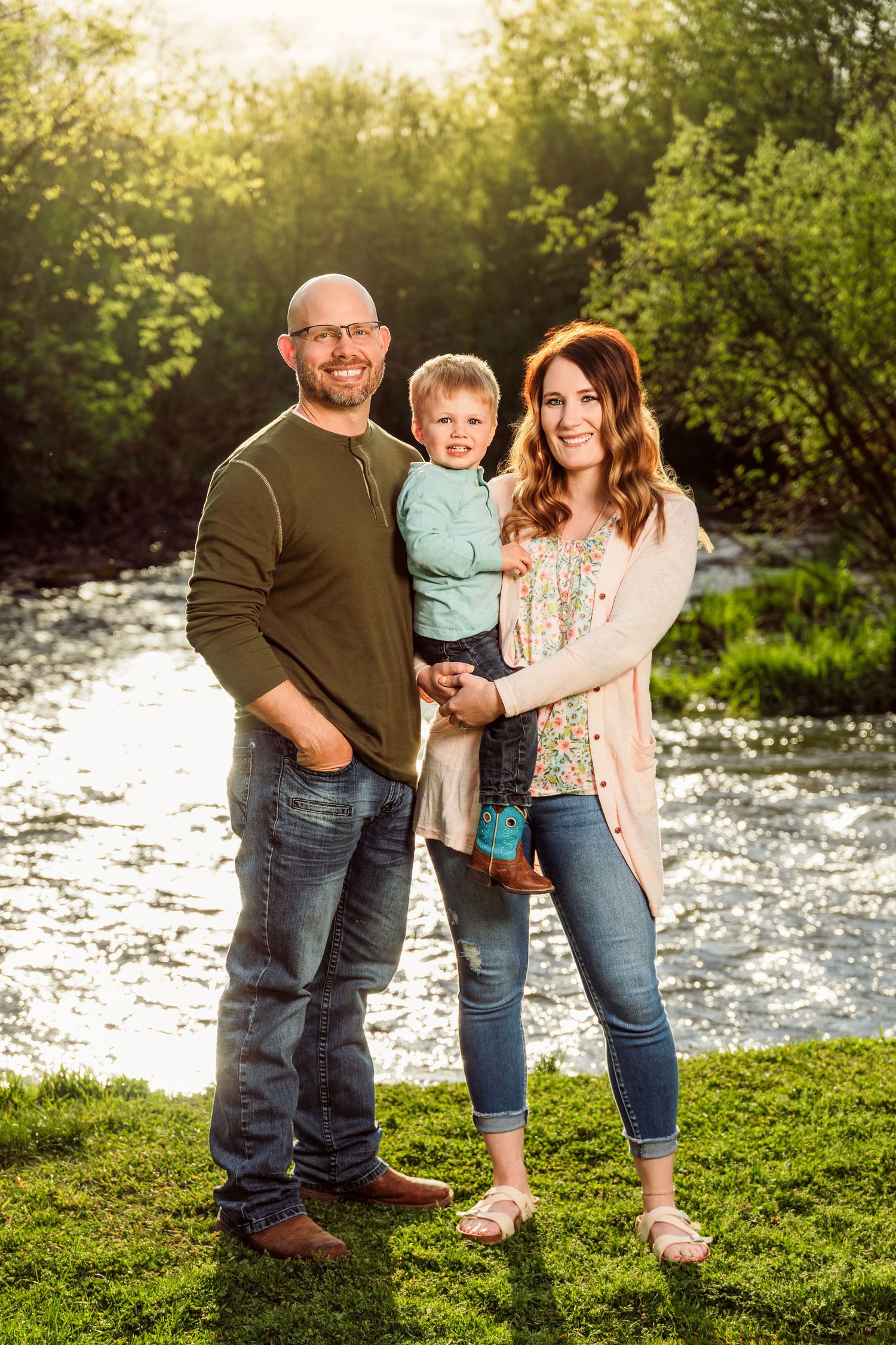 Family portrait of parents and toddler son posing by a sunlit river surrounded by lush green trees.