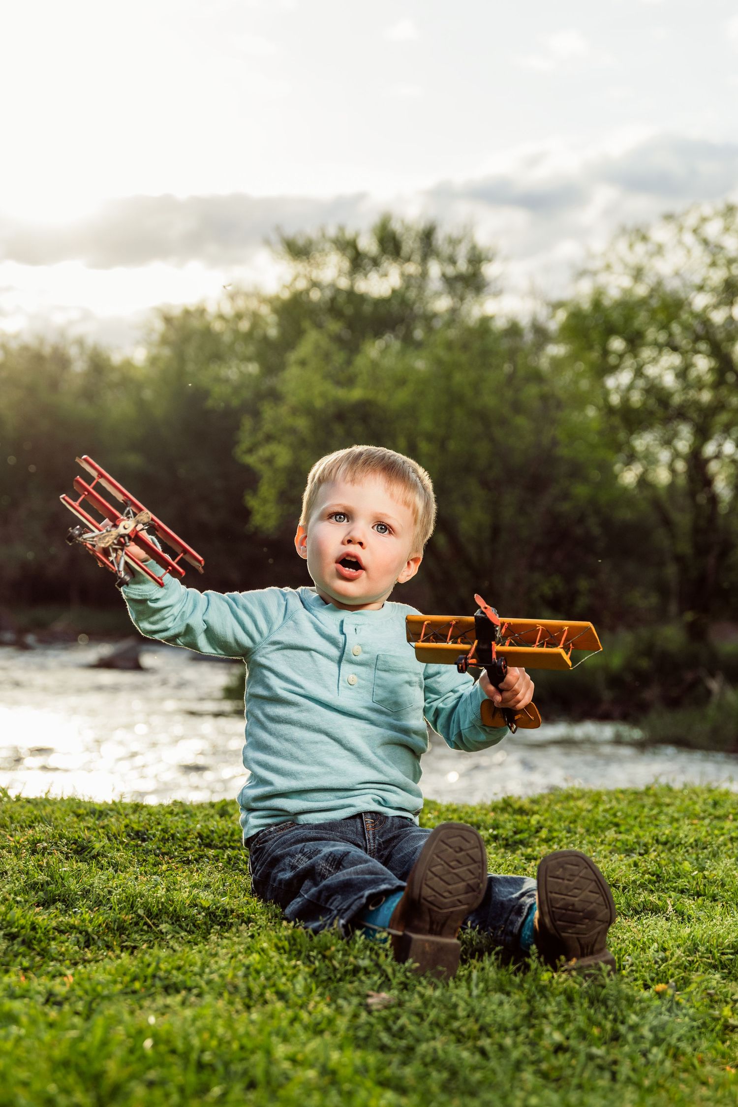 Toddler boy sitting on grass holding two toy airplanes outdoors near a river with trees in background.