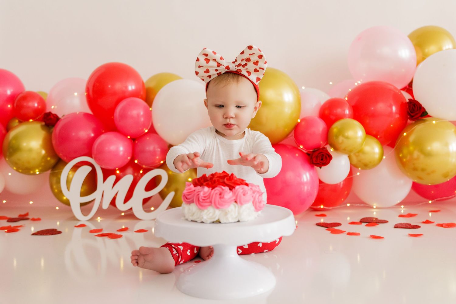 White cake stand surrounded by pink and gold balloons for a first birthday celebration.