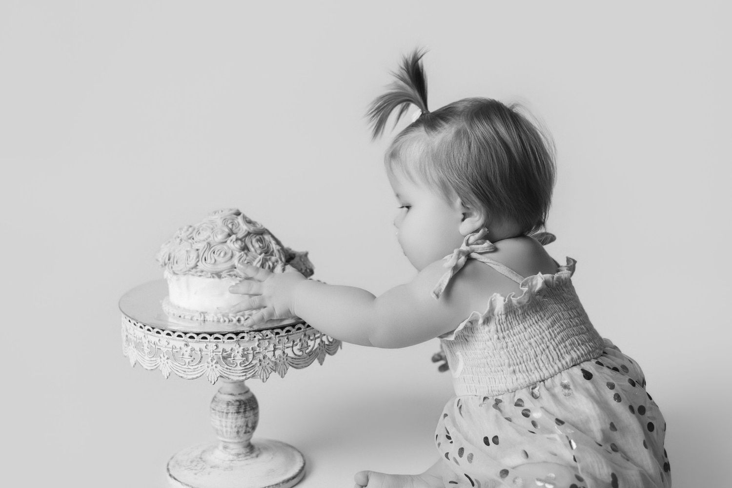 Black and white photo of baby reaching for decorated cake on ornate pedestal stand.