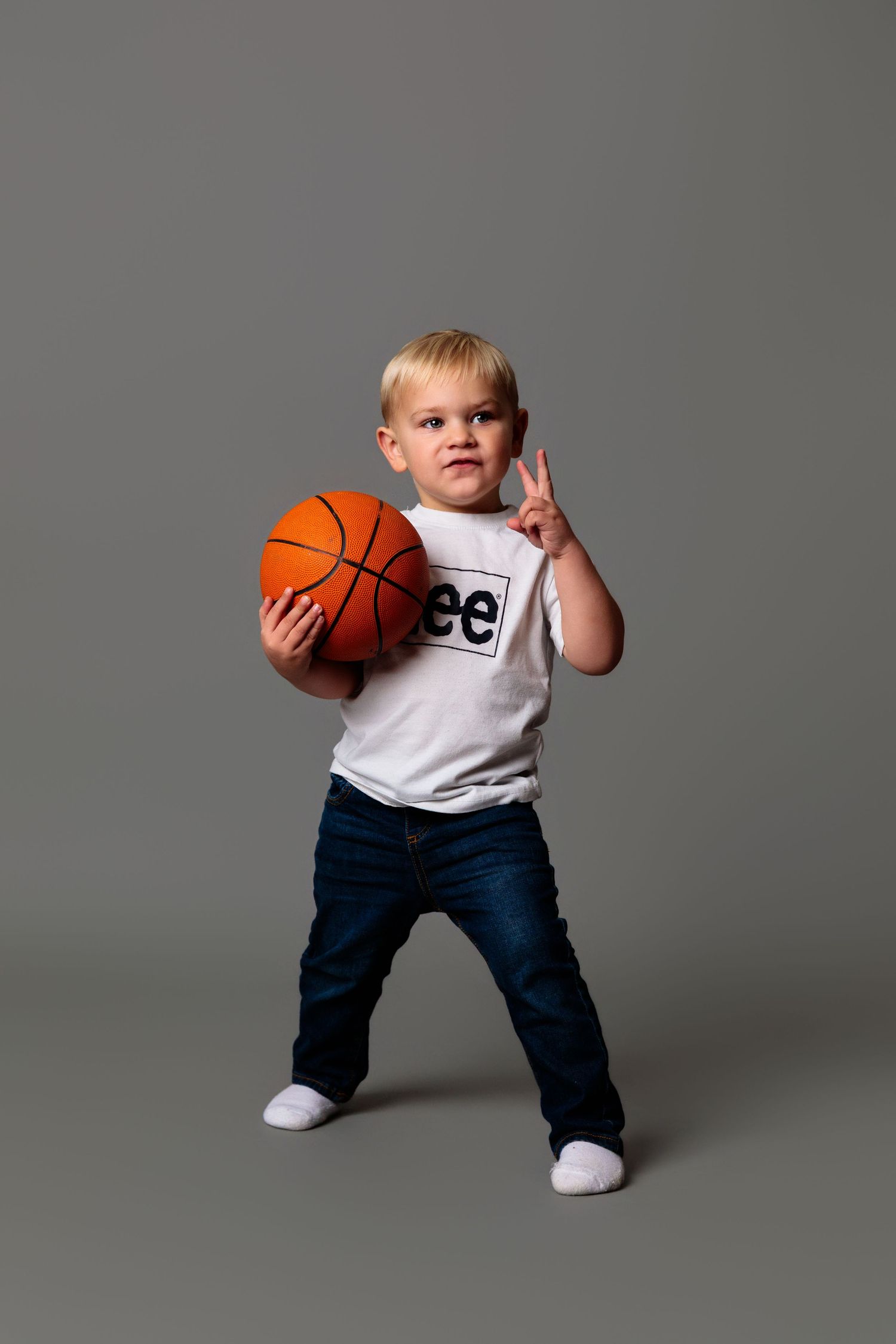 Child in white t-shirt and jeans playing with orange basketball against grey studio background.