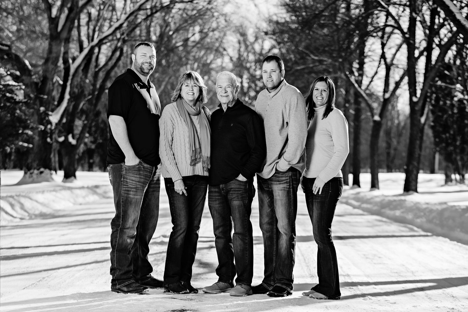 A family of five poses together on a snow-covered path surrounded by bare winter trees in black and white.
