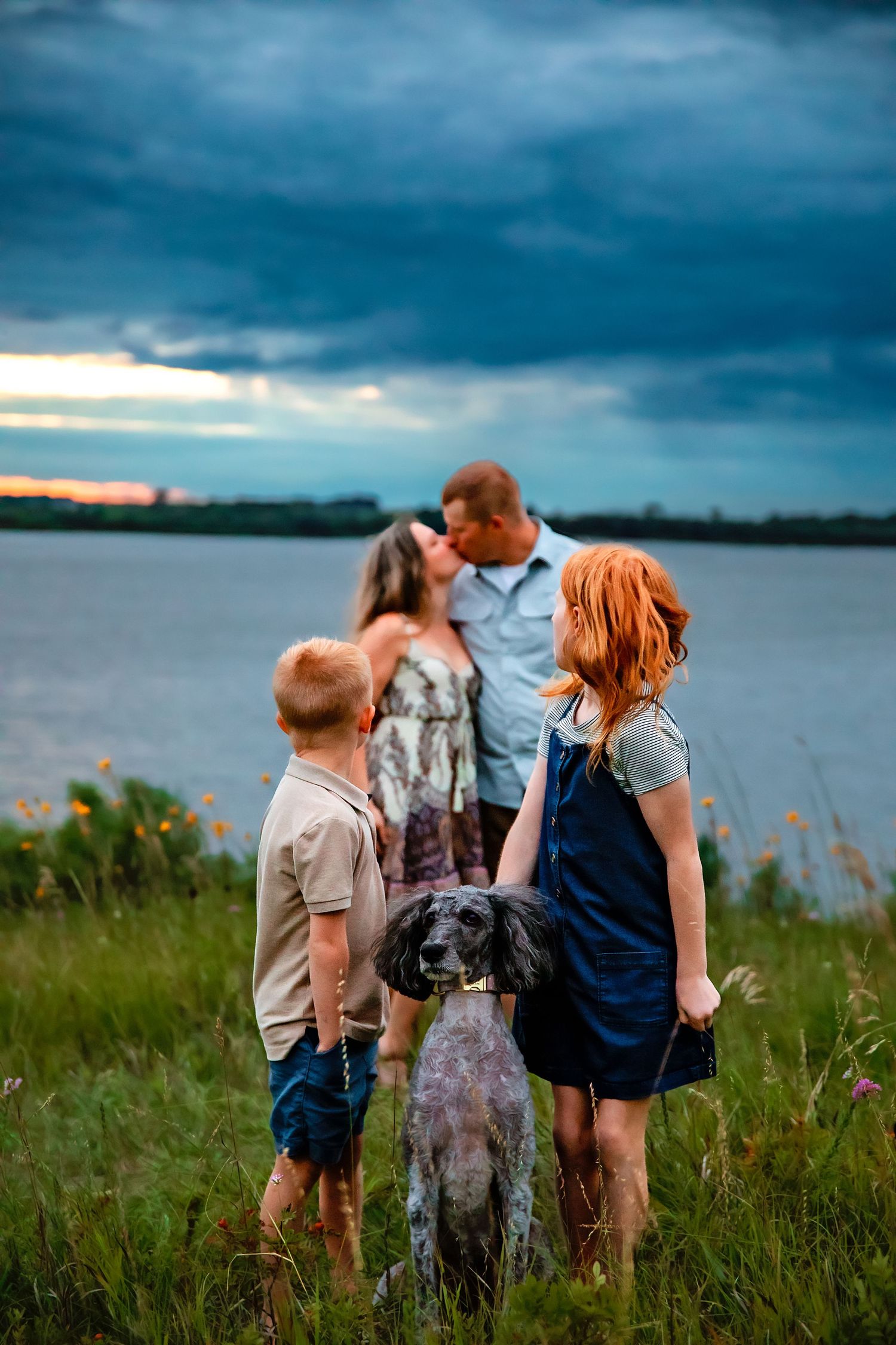 Candid family moments captured in a waterfront meadow during golden hour with dramatic storm clouds.