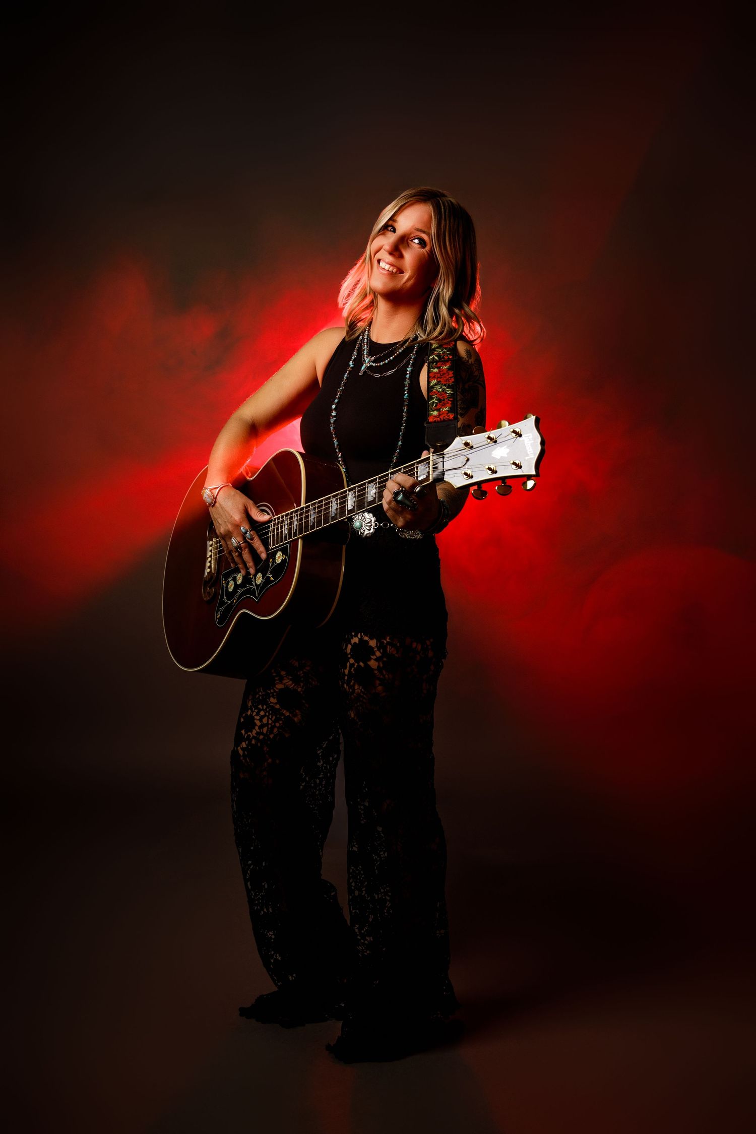 Smiling woman in black lace jumpsuit playing acoustic guitar against dramatic red smoky background.