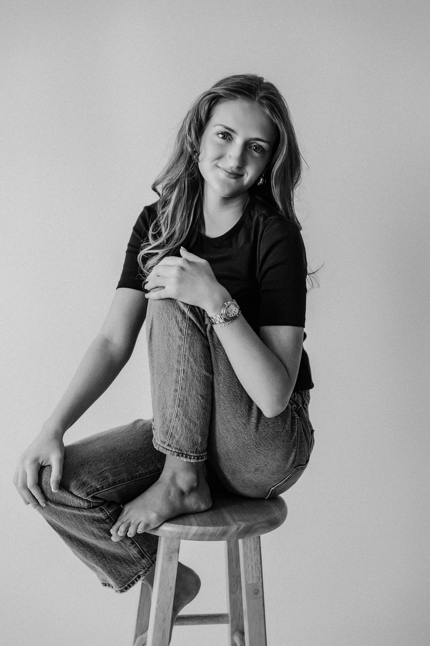 Black and white portrait of smiling young woman seated on wooden stool in black tee and jeans, barefoot.