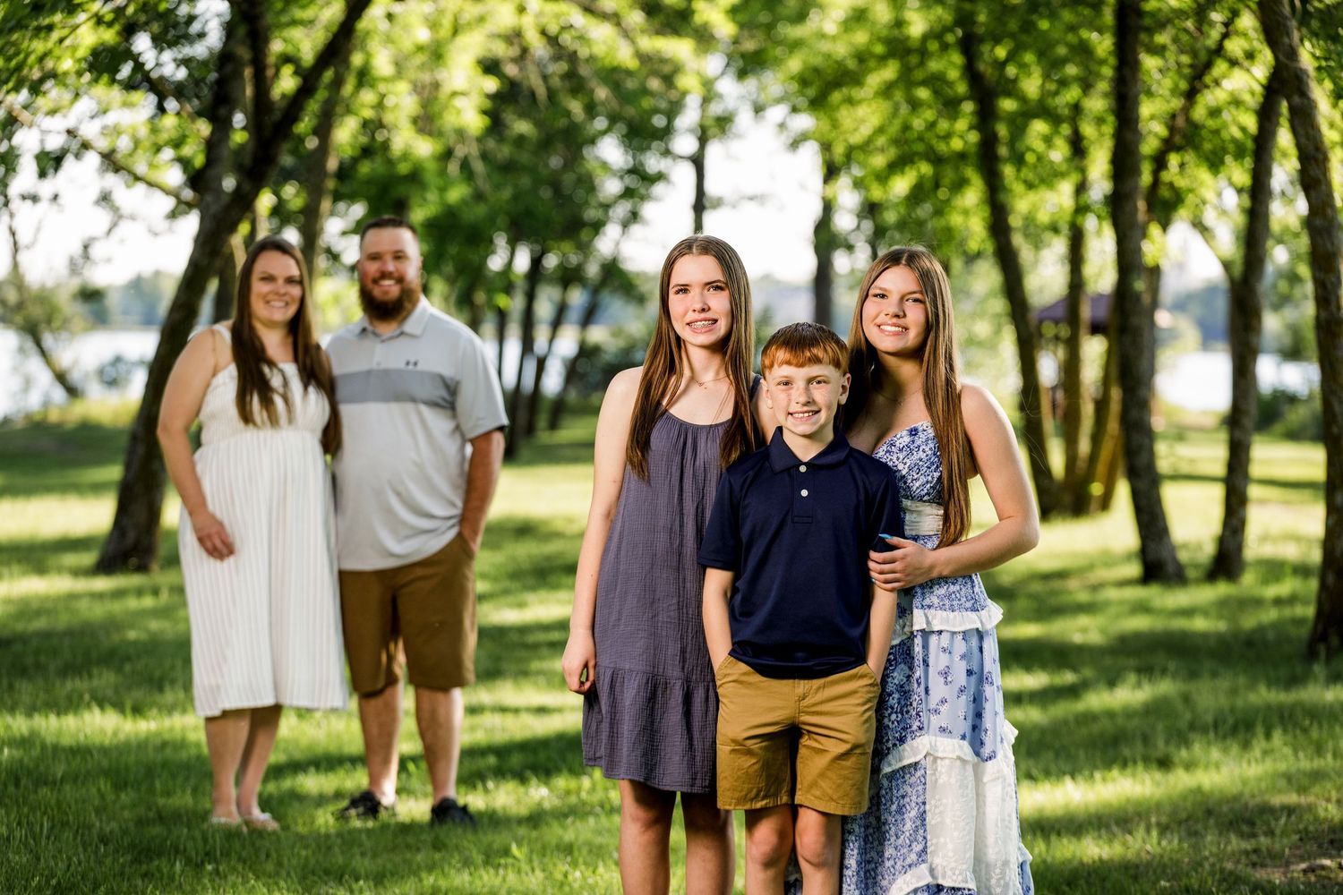 Family of five posing outdoors in a lush green park with trees and a lake in the background on a sunny day.