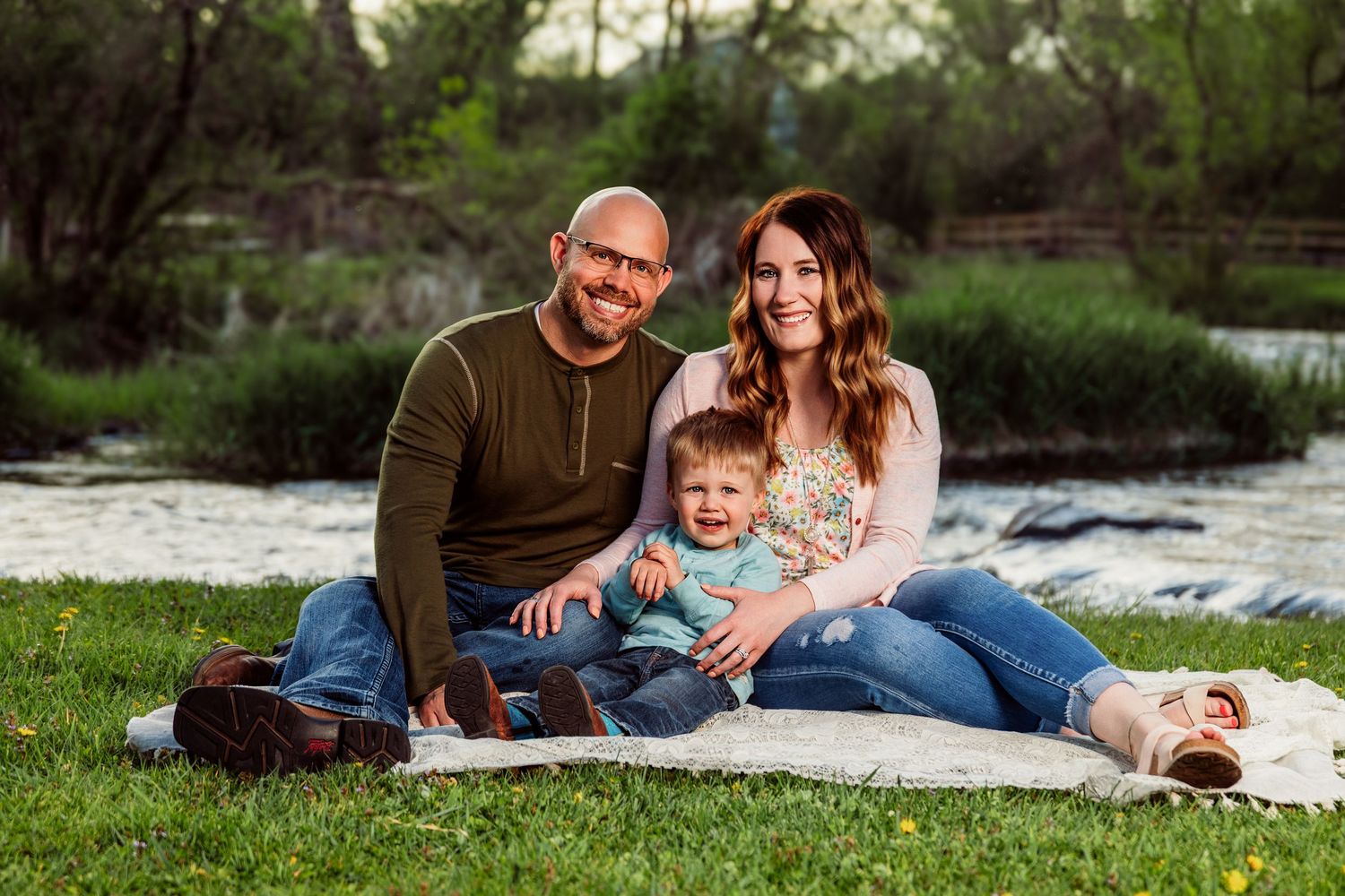 Happy family of three sitting on a blanket by a scenic river, smiling outdoors in a lush green park.