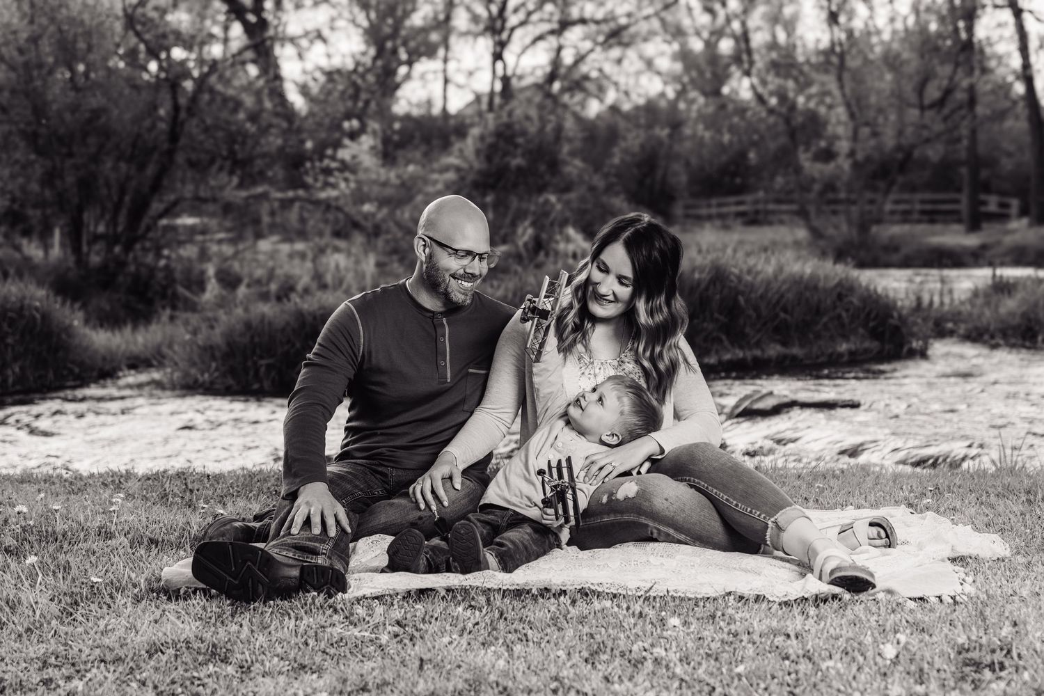 Black and white photo of a happy family sitting on a blanket outdoors, parents smiling at their newborn baby.
