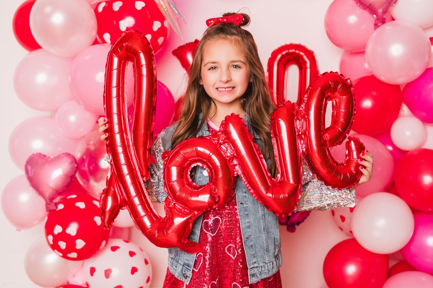 Girl holding red love balloon letters in front of pink and red Valentine's Day balloon arch backdrop.