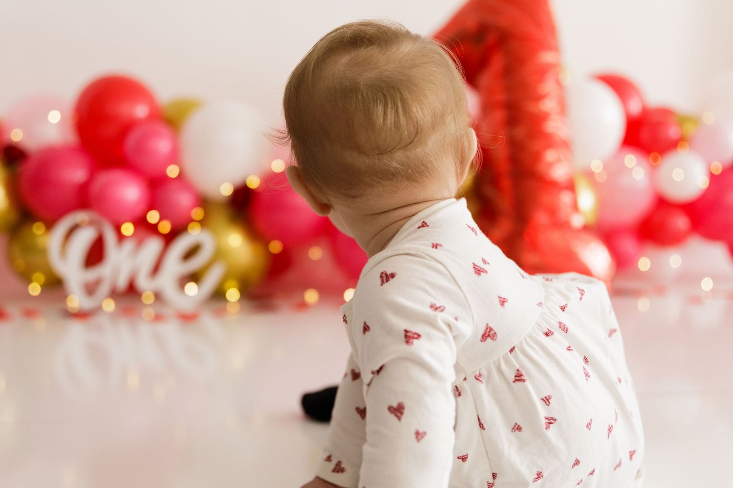 Festive first birthday setup with red and gold balloons against white backdrop for cake smash photoshoot.