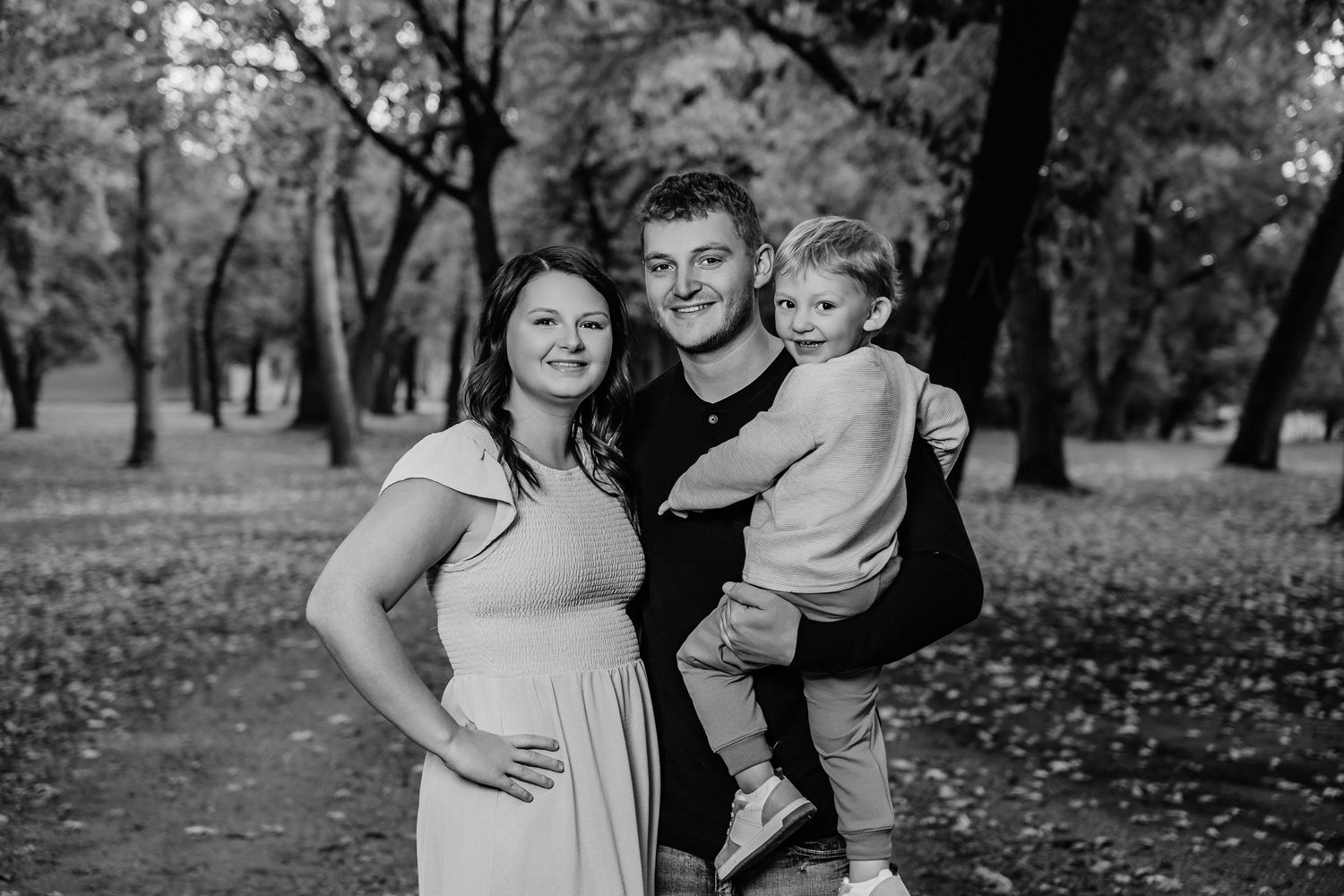 A family of three poses together on a tree-lined path covered in fallen leaves.